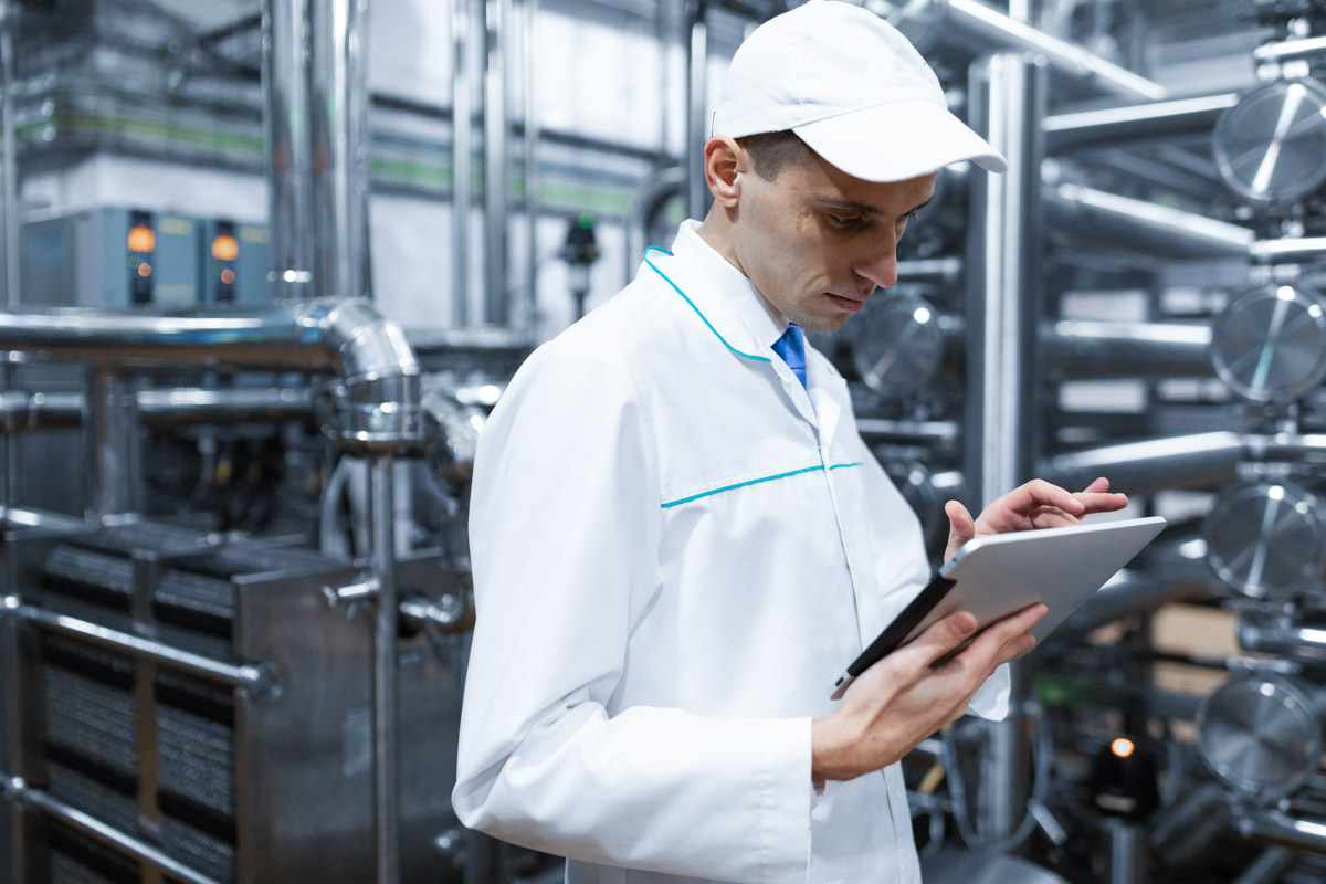 Portrait of man in a white robe and a cap standing in production department of dairy factory with grey tablet